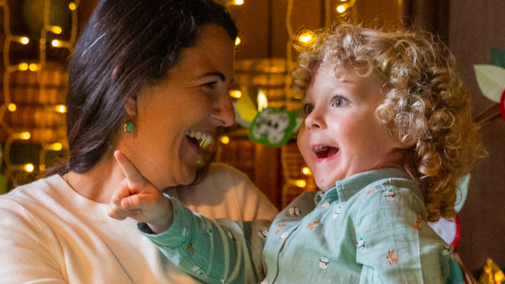 A close-up of a smiling woman and a boy pointing at Christmas decorations in Avebury Manor, against a backdrop of fairy lights.